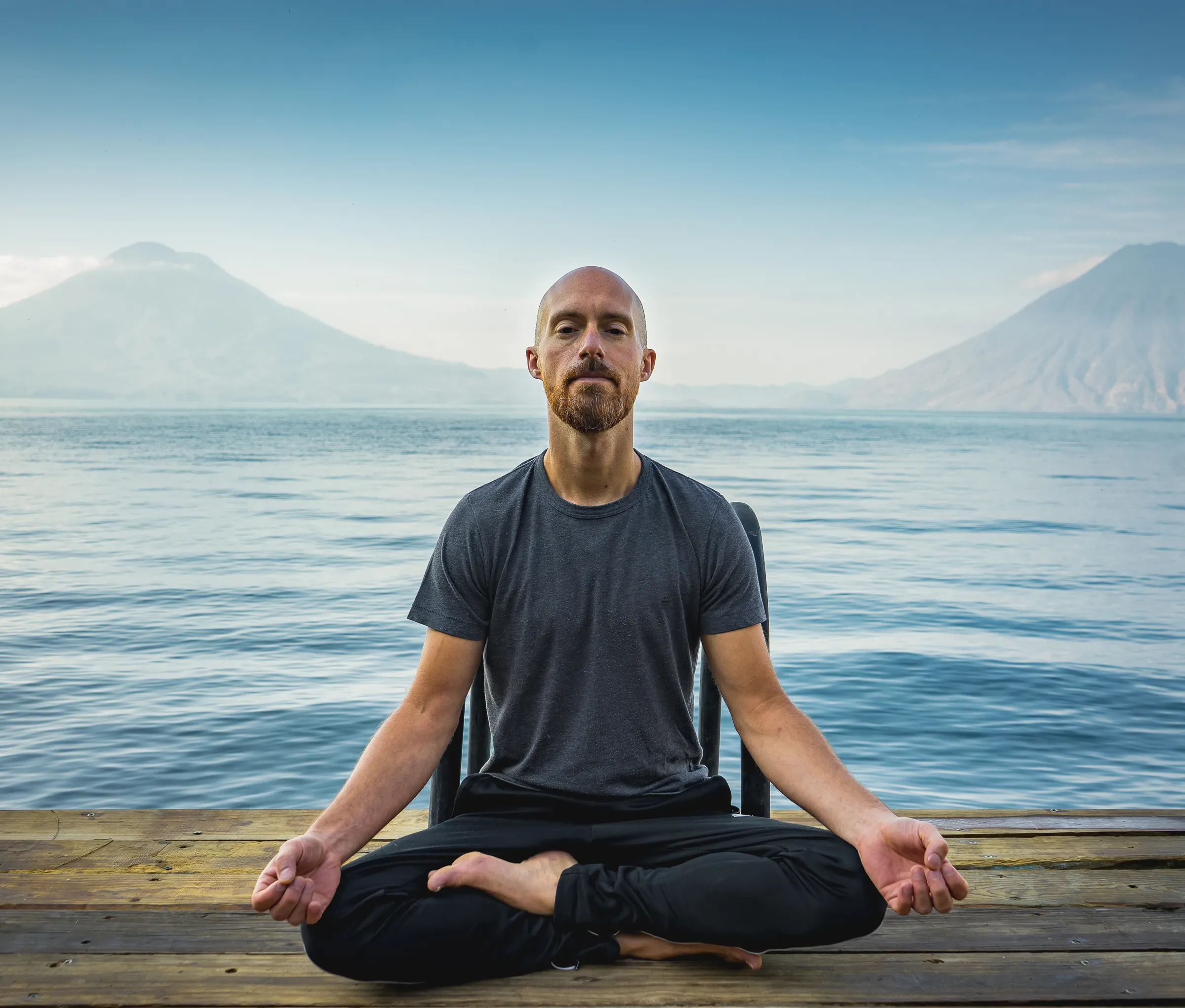 Ben sitting in meditation on a wooden pier by a calm lake, with mountains behind