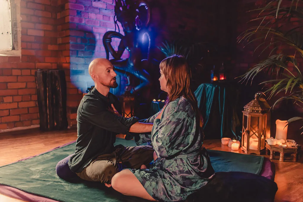 Ben and a client in a robe sitting opposite each other on a mat, talking, in a candlelit brick studio