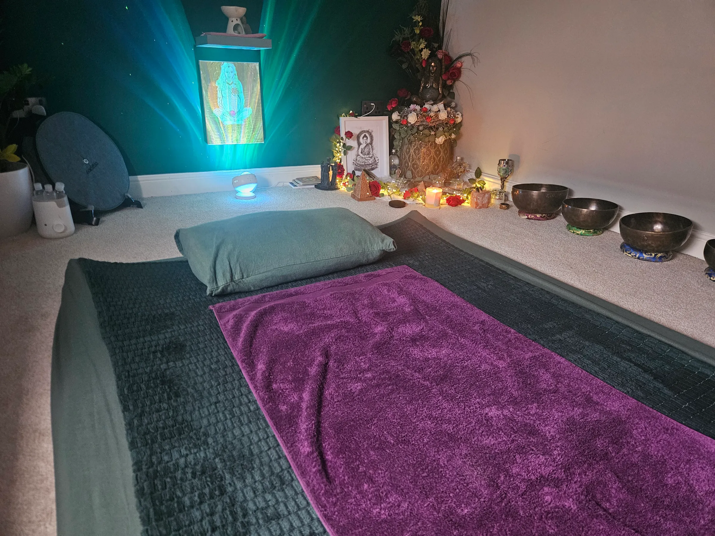 A calm treatment room with teal walls, a purple towel on a floor mattress, and singing bowls glowing in candlelight
